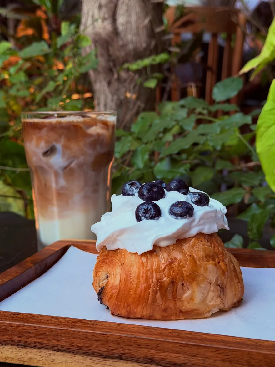 A cream-topped cruffin scattered with blueberries beside an iced latte, on a wooden board in the greenery.