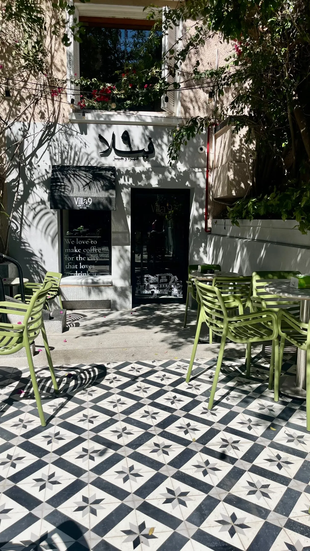 The terrace of Villa 9 in Kafr Abdo: olive-green chairs on black-and-white star tiles beneath the Villa 9 awning, with a window painted “We love to make coffee for the city that loves to drink it”.