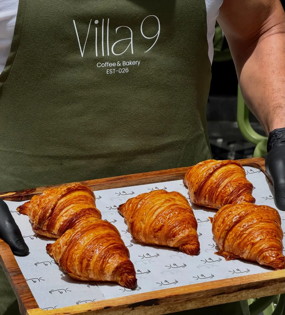 A Villa 9 apron in olive green, printed with the wordmark and EST-026, above a wooden board of butter croissants on paper printed with فيلا ٩.